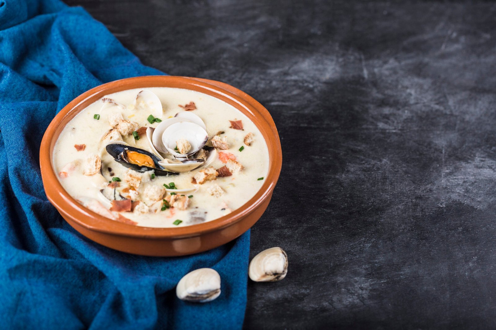 Clam chowder in a brown plate. The main ingredients are shellfish, broth, butter, potatoes and onions. New England soup also known as Boston clam chowder. On a dark gray background. Sideview. Copyspace.